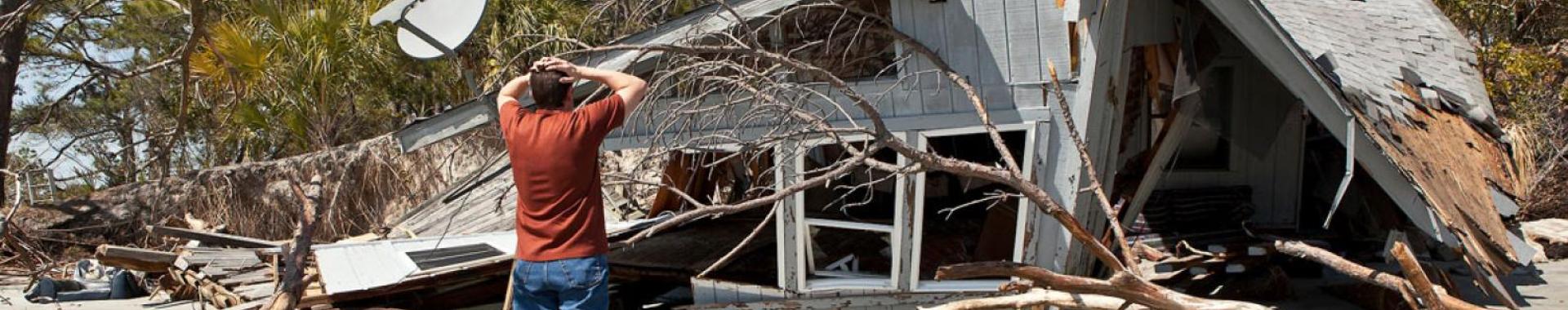 Person standing with their hands on their head in front of a house badly damaged